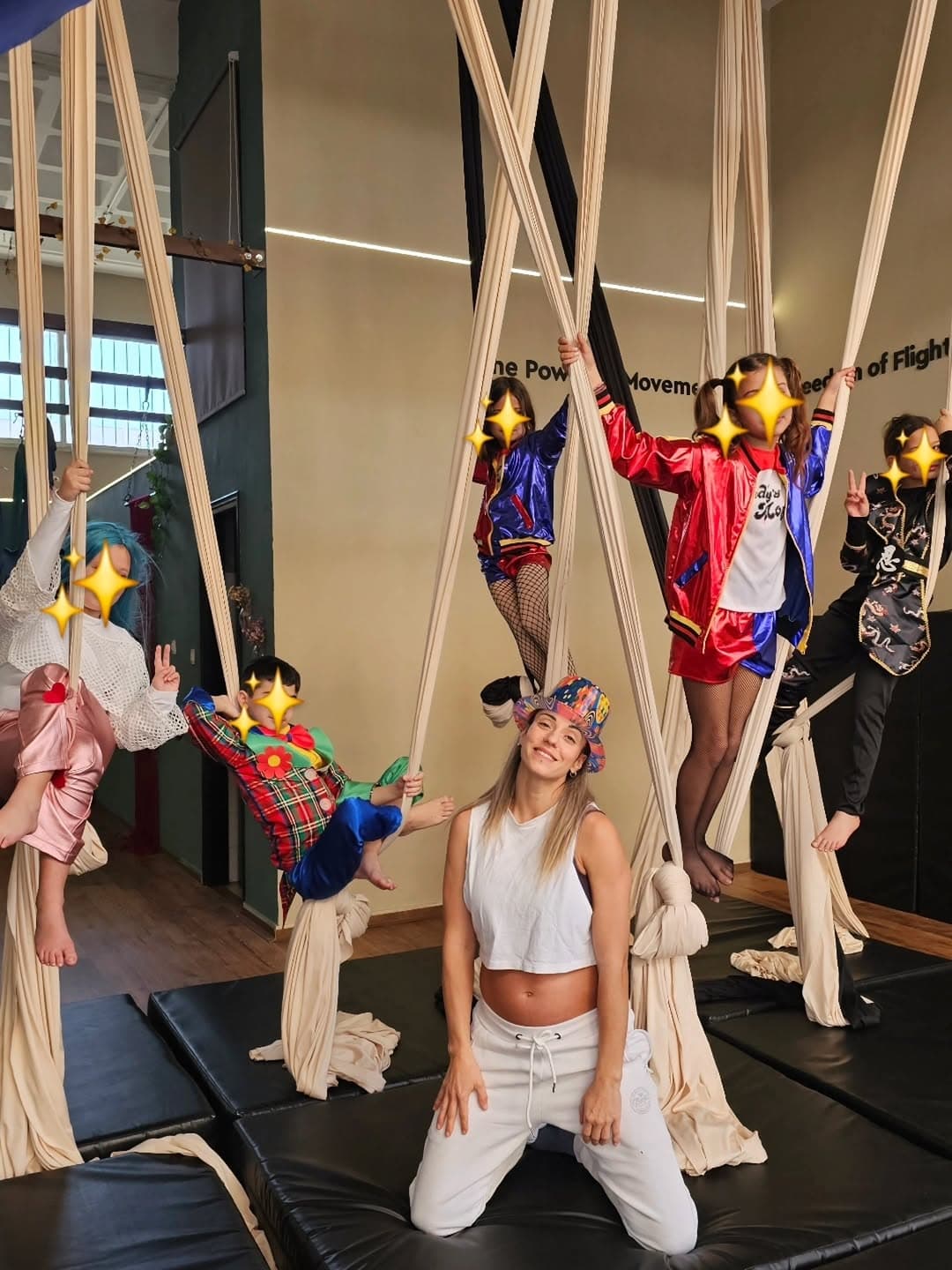 Aerial yoga gym training session with kids dressed in colorful costumes on aerial silks and a smiling instructor kneeling in front