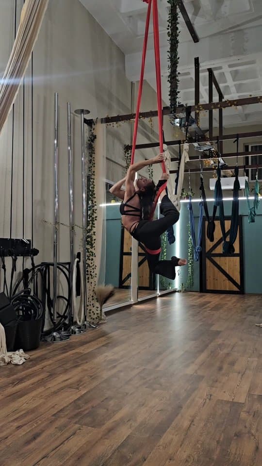 Indoor gym training center with a female athlete performing an advanced aerial silk pose using red silks, demonstrating flexibility and strength.
